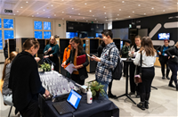 People registering for a conference in a modern indoor space. Name badges and a laptop are on the table. Attendees appear cheerful and are conversing with each other. Lockers and a large screen are visible in the background.